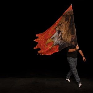 May include: A person holding a red, orange, and black flag with a lion and eagle design. The flag is torn and has a flame-like pattern.