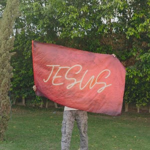 Könnte beinhalten: Eine rechteckige Flagge mit einem tiefroten, strukturierten Hintergrund und dem Wort JESUS in einer weißen, kursiven Neon-Schrift in der Mitte. Der Stoff zeigt einen Verlauf aus dunklen Rottönen und ein glattes, fließendes Erscheinungsbild.