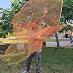 May include: A large, rectangular, sheer orange fabric prop with a yellow border, held aloft to create flowing movement. The lightweight material is translucent, allowing light to pass through its vibrant, warm-toned surface while it billows in the air.
