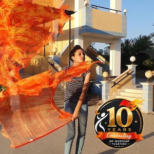 May include: Two large, lightweight worship flags featuring a vibrant, swirling flame pattern in shades of orange, red, and yellow. The fabric is translucent and fluid, creating dynamic movement. A circular logo with the text 10 YEARS CELEBRATING OF WORSHIP TOGETHER is visible in the lower right corner.