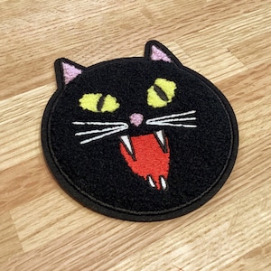 May include: Black cat patch with yellow eyes, pink nose and ears, and a red open mouth with white fangs. The patch is round with a black border and is on a wooden surface.