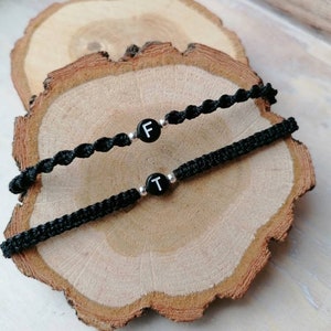 Könnte beinhalten: Zwei schwarze Freundschaftsarmbänder mit silbernen Perlen und schwarzen Buchstabenanhängern. Die Anhänger buchstabieren die Buchstaben "F" und "T".