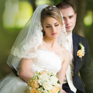 Pu&ograve; includere: Una sposa e uno sposo posano per un ritratto di matrimonio. La sposa indossa un abito da sposa bianco con un velo e tiene un bouquet di fiori bianchi e pesca. Lo sposo indossa un abito scuro con una camicia bianca e un boutonni&egrave;re.