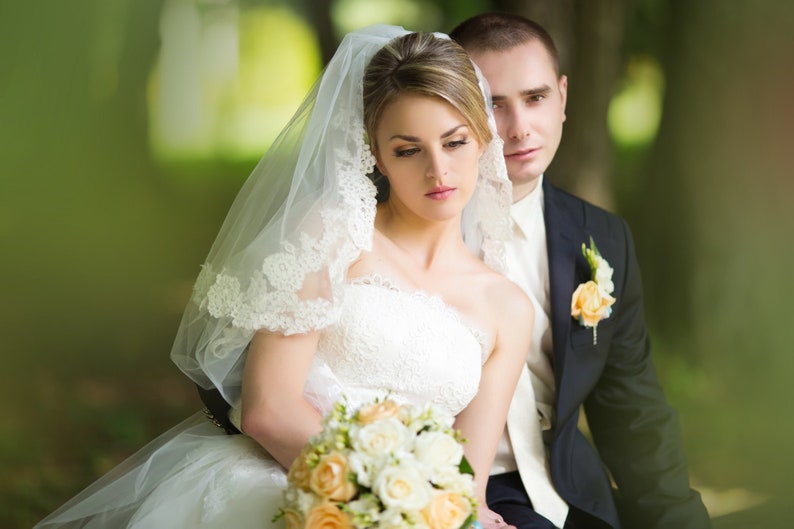 Pu&ograve; includere: Una sposa e uno sposo posano per un ritratto di matrimonio. La sposa indossa un abito da sposa bianco con un velo e tiene un bouquet di fiori bianchi e pesca. Lo sposo indossa un abito scuro con una camicia bianca e un boutonni&egrave;re.