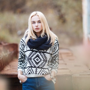 Pu&ograve; includere: Una giovane donna con lunghi capelli biondi indossa un maglione a motivi bianchi e neri con una sciarpa blu scuro. &Egrave; in piedi davanti a una superficie di metallo arrugginito.