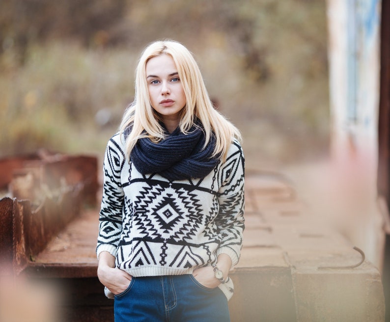 Pu&ograve; includere: Una giovane donna con lunghi capelli biondi indossa un maglione a motivi bianchi e neri con una sciarpa blu scuro. &Egrave; in piedi davanti a una superficie di metallo arrugginito.