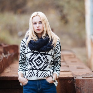 Pu&ograve; includere: Una donna con i capelli biondi indossa un maglione a motivi bianchi e neri con una sciarpa blu scuro. &Egrave; in piedi davanti a una superficie di metallo arrugginito.