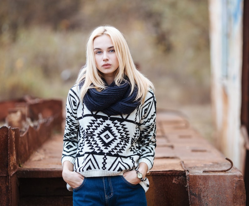 Pu&ograve; includere: Una donna con i capelli biondi indossa un maglione a motivi bianchi e neri con una sciarpa blu scuro. &Egrave; in piedi davanti a una superficie di metallo arrugginito.