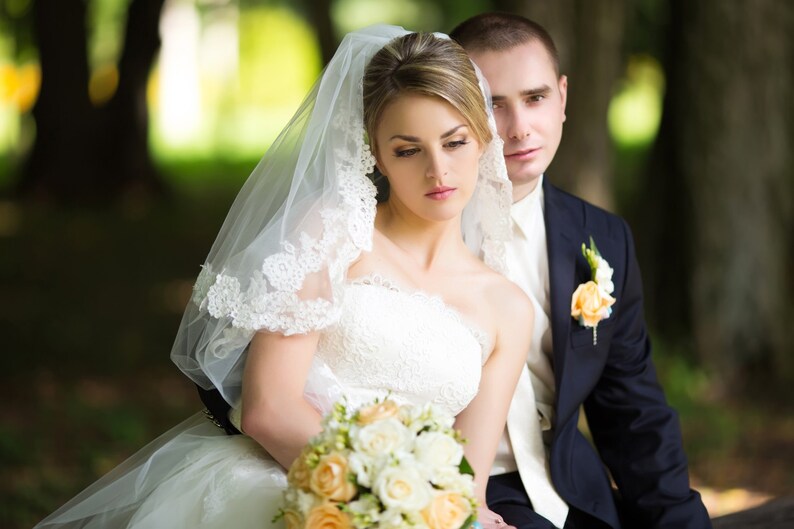 Pu&ograve; includere: Un ritratto di matrimonio di una sposa e uno sposo in un parco. La sposa indossa un abito da sposa bianco con un velo e un bouquet di fiori bianchi e gialli. Lo sposo indossa un abito scuro e una camicia bianca con una cravatta.
