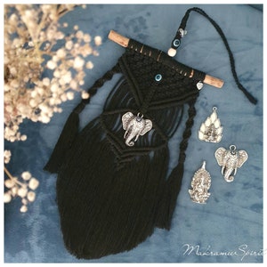 Könnte beinhalten: Schwarzer Makramee-Wandbehang mit einem silbernen Elefanten-Anhänger, zwei silbernen Anhängern einer Gottheit und einem blauen Glücksbringer-Anhänger. Der Wandbehang ist an einem Holzstab befestigt.