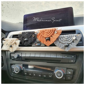 Könnte beinhalten: Vier Makramee-Autolufterfrischer, die an den Lüftungsschlitzen eines Autos hängen. Die Lufterfrischer sind in den Farben Beige, Schwarz, Orange und Grau. Jeder hat einen kleinen Holzstab und eine schwarze Perle, die daran hängen. Der Text "Makramee Spirit" ist auf dem Bildschirm im Hintergrund sichtbar.