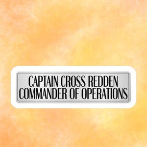 May include: A rectangular sign with the text "CAPTAIN CROSS REDDEN COMMANDER OF OPERATIONS" in black letters. The sign has a silver metallic appearance and a white border, set against a watercolor-style orange and yellow background.