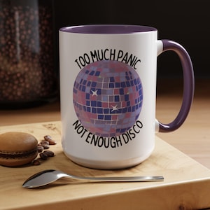 May include: White ceramic mug with a purple interior and handle. The mug features a disco ball graphic in shades of blue, pink, and purple, with the text "TOO MUCH PANIC NOT ENOUGH DISCO" in black. The mug is on a wooden surface.