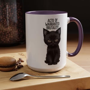 Puede incluir: Taza de cerámica blanca con interior y asa morados. La taza presenta una ilustración de un gato negro y el texto "ACTS OF WARRANTED BRUTALITY". La taza está sobre una superficie de madera con un macaron, granos de café y una cuchara.