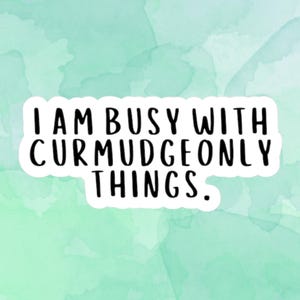 May include: A sticker with the text "I AM BUSY WITH CURMUDGEONLY THINGS" in a black, bold font. The text is on a white background, set against a watercolor wash of light green and blue.