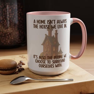 Könnte beinhalten: Weißer Keramikbecher mit rosa Innenraum und Henkel. Der Becher zeigt eine braune Illustration eines Hauses und den Text: „A HOME ISN'T ALWAYS THE HOUSE WE LIVE IN. IT'S ALSO THE PEOPLE WE CHOOSE TO SURROUND OURSELVES WITH.“
