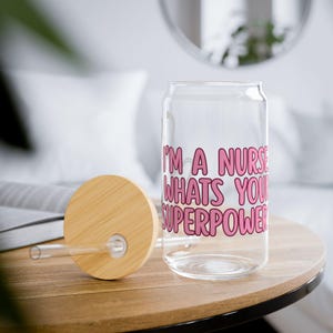 May include: A clear glass can-shaped drinking glass with a bamboo lid and straw. The glass has pink text that reads "I'M A NURSE WHAT'S YOUR SUPERPOWER". The glass is on a wooden table.