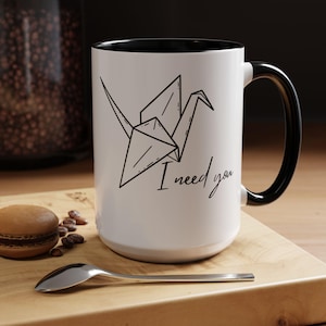 Op de afbeelding: Witte keramische mok met een zwart interieur en handvat. De mok heeft een zwarte lijntekening van een origami kraanvogel en de handgeschreven tekst "I need you". Een lepel en een macaron liggen op het houten oppervlak.