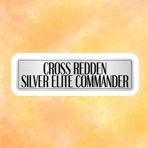 May include: A rectangular sign with rounded corners, featuring the text "CROSS REDDEN SILVER ELITE COMMANDER" in black capital letters. The sign has a silver metallic appearance and is set against a watercolor-style orange and yellow background.