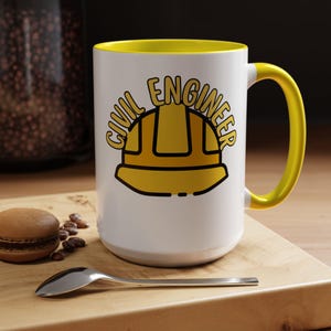 Puede incluir: Taza de cerámica blanca con interior y asa amarillos. La taza presenta las palabras "CIVIL ENGINEER" en amarillo, junto con un gráfico de casco amarillo. La taza está sobre una superficie de madera con una cuchara, granos de café y un macaron.