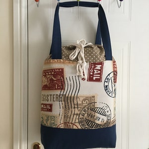 May include: A beige tote bag with navy blue handles and a bottom. The bag is made of fabric with a vintage postage stamp design. The fabric has a white polka dot pattern. The bag has a white rope handle.