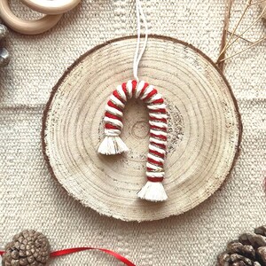 Könnte beinhalten: Handgefertigter Zuckerstangen-Ornament mit rot-weißen Streifen, goldfarbenen Fäden und weißen Quasten. Der Ornament hängt an einer weißen Schnur und wird auf einer Holzscheibe mit neutralem Hintergrund präsentiert. Weitere Dekorationen sind Tannenzapfen und Holzringe.