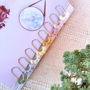 Può includere: Un set di sette orecchini in metallo dorato a forma di U, ognuno caratterizzato da una piccola sezione di cordino intrecciato di colore diverso. I colori includono bianco, rosa chiaro, rosa antico, giallo senape, crema, azzurro e verde salvia.