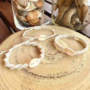 Può includere: Tre braccialetti fatti a mano realizzati con cordino in macramè color bianco sporco intrecciato in motivi complessi, ognuno centrato con una singola conchiglia di ciprea liscia color crema. I braccialetti presentano chiusure regolabili.