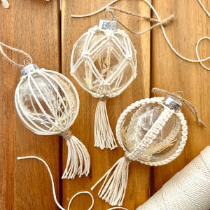 Può includere: Tre palline di vetro trasparente con composizioni floreali essiccate all'interno, avvolte in macramè e decorate con nappe. Le palline sono appese ad anelli di spago. Sono visibili anche una bobina di cordoncino bianco e dello spago sciolto.