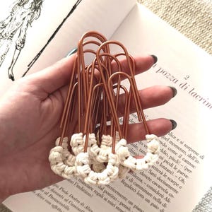 May include: A set of large copper-colored metal paper clips, each featuring a decorative knot of off-white macrame cord tied around the bottom loop. The clips are arranged in a cluster, showcasing the metallic finish and the textured fiber knots.