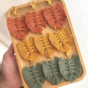 Peut inclure: Une collection de porte-clés feuilles en macramé faits à la main, dans des tons d'orange, de jaune et de vert. Chaque porte-clés est doté d'un fermoir doré et est présenté sur un plateau en bois. Les porte-clés sont en forme de feuille.
