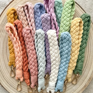 Puede incluir: Una colección de llaveros de pulsera de macramé trenzado en varios colores sólidos, incluyendo mostaza, melocotón, terracota, rosa, lavanda, azul claro, blanco, verde salvia y verde oscuro, cada uno con un mosquetón de metal dorado en la base.