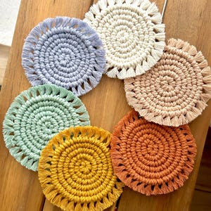 Op de afbeelding: Zes ronde gehaakte onderzetters met franjes, in verschillende kleuren, waaronder lichtblauw, wit, lichtroze, groen, geel en oranje. De onderzetters zijn in een cirkel op een houten oppervlak gerangschikt.