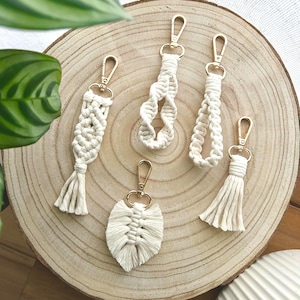 Peut inclure: Cinq porte-clés macramé faits à la main de différentes formes, dont une feuille, des pompons et des motifs tressés. Chaque porte-clés est doté d'un fermoir doré. Les porte-clés sont présentés sur une tranche de bois clair, avec un feuillage vert en arrière-plan.