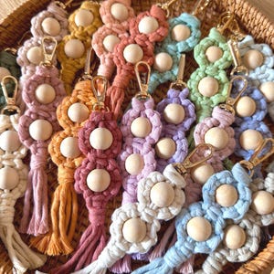 Puede incluir: Una colección de llaveros de macramé en varios colores, cada uno con dos cuentas de madera redondas y un mosquetón de metal dorado. Los diseños de cordón de algodón anudado incluyen tonos de rosa, amarillo, naranja, azul, verde y blanco, y cada llavero termina en una borla corta y deshilachada.