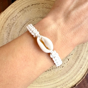 Può includere: Un braccialetto in macramè bianco con una conchiglia naturale al centro. Il braccialetto è realizzato con un cordoncino bianco intrecciato e viene mostrato su un polso. La conchiglia è di colore crema chiaro.