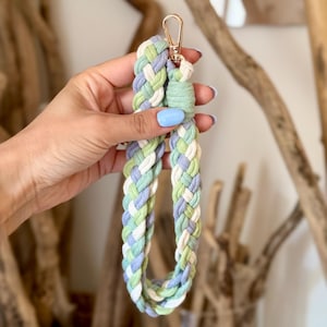 May include: A braided wristlet keychain made of soft fabric cord in pastel shades of sage green, lavender, and white. The braided loop is secured with a matching sage green cord wrap at the top and features a gold-toned metal lobster clasp.