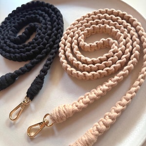 May include: Two macrame bag straps in black and beige, coiled on a white plate. Each strap has a gold-colored clasp. The black strap is coiled on the left, while the beige strap is coiled on the right.