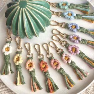 May include: A selection of handmade flower keyrings in various colours, including white, yellow, peach, orange, pink, blue, and purple. Each keyring features a gold-coloured clasp and a tassel made of green, yellow, and peach-coloured threads. The keyrings are arranged on a white plate.