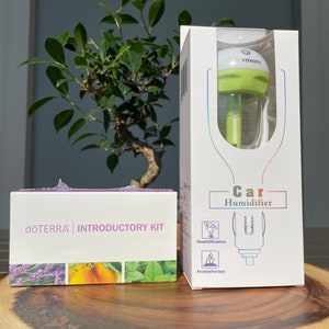 May include: A white box with a green and white car humidifier inside. The box says "Car Humidifier" and has icons for humidification and aromatherapy. The box is next to a white box with the text "dōTERRA INTRODUCTORY KIT" and images of lavender, orange, and mint.