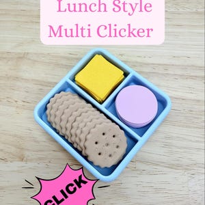 May include: A light blue divided tray containing a stack of tan scalloped round crackers, a stack of yellow square crackers, and a stack of lavender circular discs. The text "Lunch Style Multi Clicker" appears above, and a pink starburst says "CLICK".