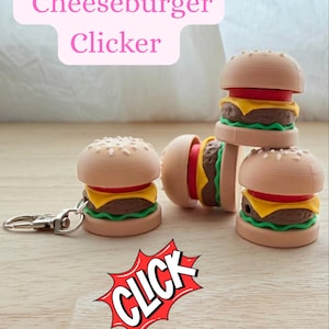 Op de afbeelding: Een verzameling miniatuur cheeseburger clickers. Elke clicker heeft een broodje, een patty, kaas en een plakje tomaat. Eén clicker is bevestigd aan een sleutelhanger. De afbeelding bevat de tekst "Cheeseburger Clicker" en een stripboekachtige "CLICK" graphic.