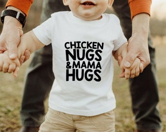 May include: A white toddler t-shirt with black text that reads "Chicken Nugs & Mama Hugs". A toddler is holding hands with an adult while wearing the shirt.