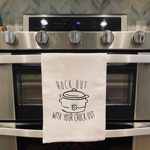 May include: White kitchen towel with black text that reads "Rock out with your crock out". A black and white illustration of a slow cooker is above the text.