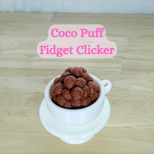 May include: A white ceramic teacup and saucer set filled with a cluster of brown, spherical objects, resembling cereal. The text "Coco Puff Fidget Clicker" is displayed above the cup in pink letters.