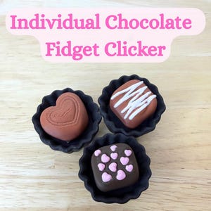 May include: Three chocolate-shaped fidget clickers in black paper cups. One is heart-shaped, one is square with pink hearts, and one is square with white lines. The text "Individual Chocolate Fidget Clicker" is above the clickers.