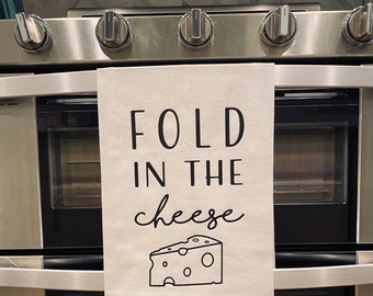 May include: A white kitchen towel with black text that reads "FOLD IN THE cheese" and a black and white illustration of a block of cheese.