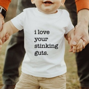 May include: A white t-shirt with the text "i love your stinking guts." printed on it. A child is wearing the shirt and holding hands with an adult.