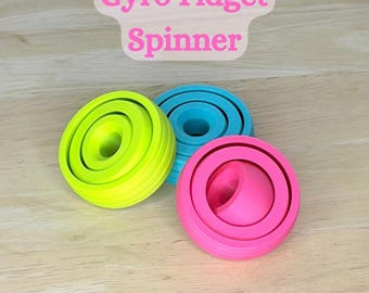 Peut inclure: Trois toupies fidget gyro colorées en vert fluo, bleu sarcelle et rose. Le texte "Gyro Fidget Spinner" est affiché au-dessus des toupies. Les toupies sont sur une surface en bois clair.