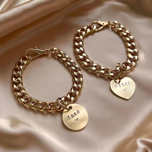 May include: Two gold-toned metal chain bracelets featuring lobster claw clasps. One bracelet has a round pendant engraved with the name Luna and a small heart, while the other has a heart-shaped pendant engraved with the name Riley and a heart.